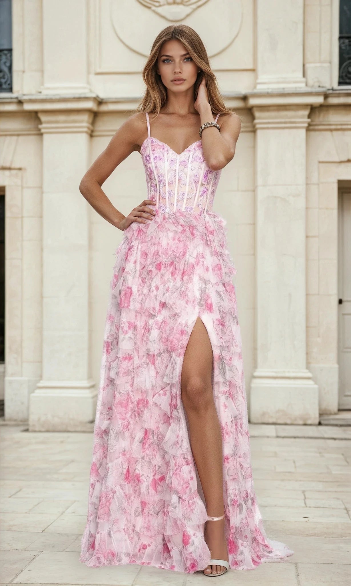 A woman stands outdoors by a stone building in the Ruffled Tiered Pink Floral-Print Prom Dress L526 (Pink Print) with a thigh-high slit, paired with light heels and minimal jewelry.